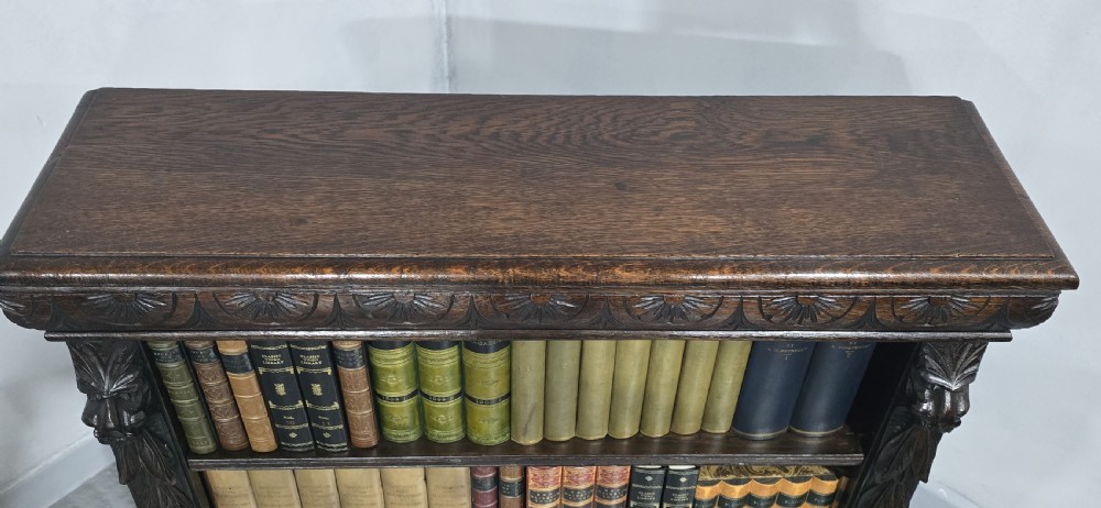 A dark carved oak open bookcase featuring four shelves filled with leather-bound books, flanked by carved foliate pilasters and topped with a decorative frieze. View VIII