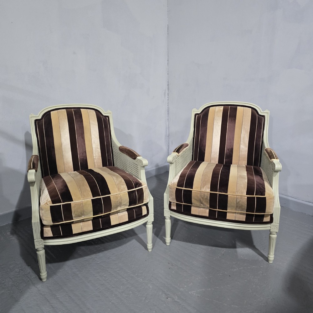 A pair of Louis XVI style French bergere armchairs featuring off-white painted wooden frames, caned sides, fluted legs, and striped brown velvet cushioned seats.