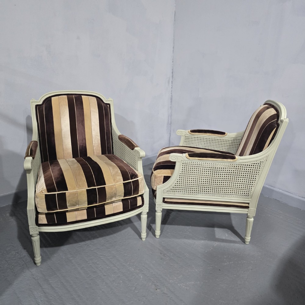 A pair of Louis XVI style French bergere armchairs featuring off-white painted wooden frames, caned sides, fluted legs, and striped brown velvet cushioned seats. View II