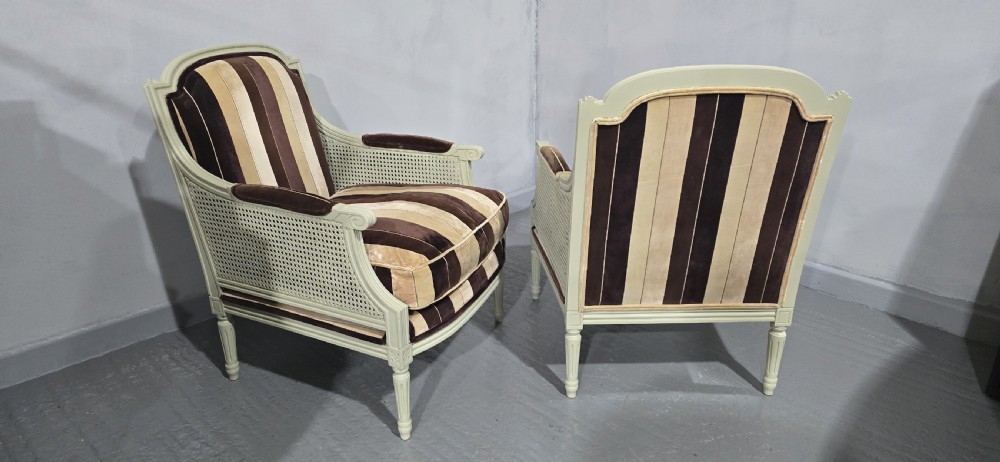A pair of Louis XVI style French bergere armchairs featuring off-white painted wooden frames, caned sides, fluted legs, and striped brown velvet cushioned seats. View V