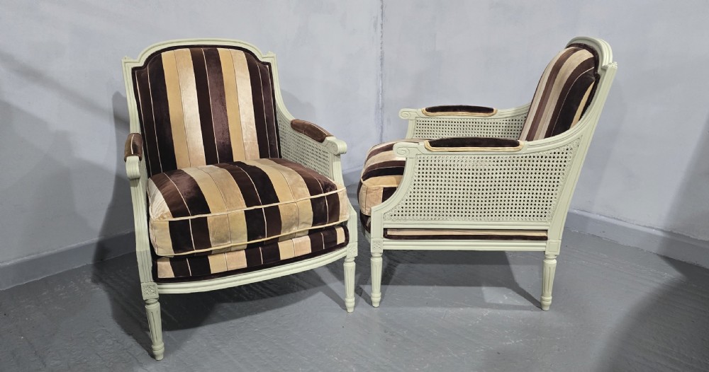 A pair of Louis XVI style French bergere armchairs featuring off-white painted wooden frames, caned sides, fluted legs, and striped brown velvet cushioned seats. View VI