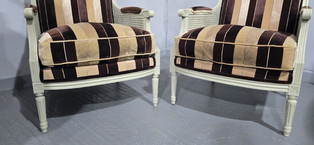 A pair of Louis XVI style French bergere armchairs featuring off-white painted wooden frames, caned sides, fluted legs, and striped brown velvet cushioned seats. View X