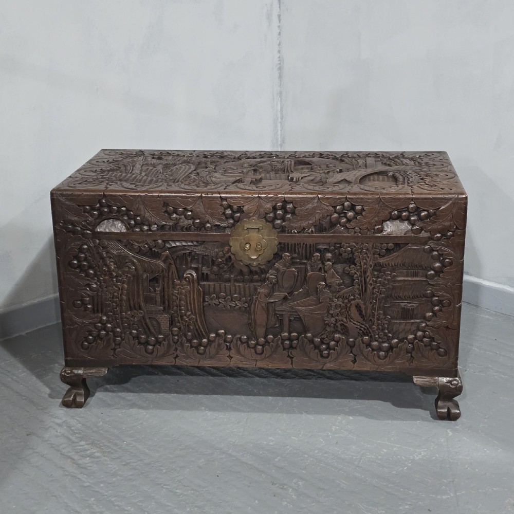 A rectangular dark timber storage chest featuring detailed relief carvings of figures and landscapes on all sides with a central brass clasp.
