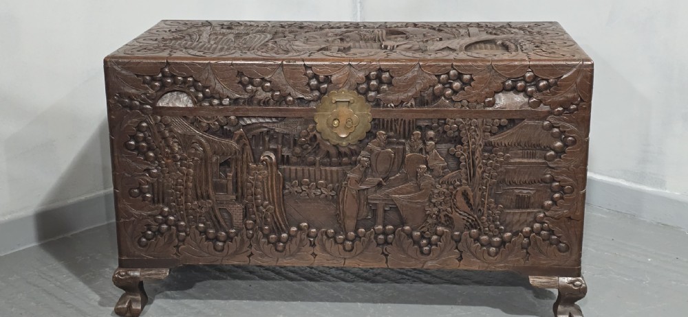 A rectangular dark timber storage chest featuring detailed relief carvings of figures and landscapes on all sides with a central brass clasp. View II