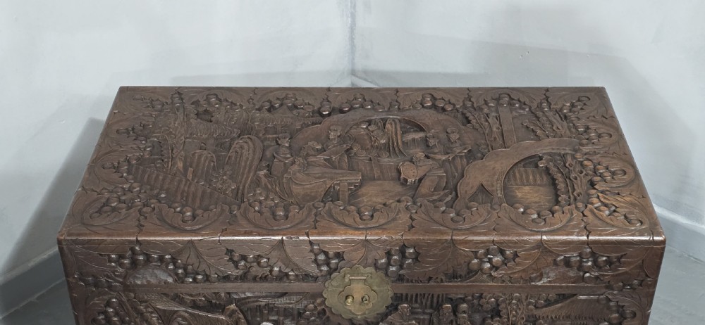 A rectangular dark timber storage chest featuring detailed relief carvings of figures and landscapes on all sides with a central brass clasp. View III