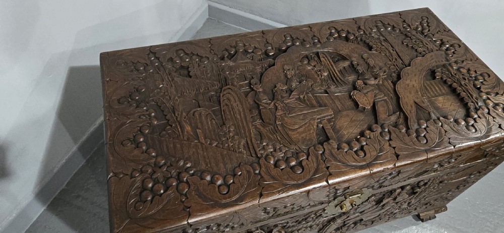 A rectangular dark timber storage chest featuring detailed relief carvings of figures and landscapes on all sides with a central brass clasp. View VI