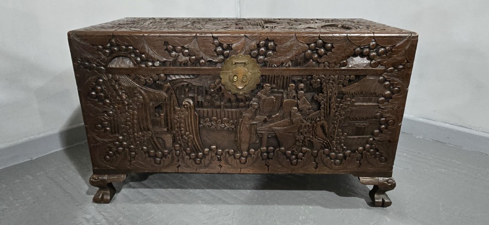 A rectangular dark timber storage chest featuring detailed relief carvings of figures and landscapes on all sides with a central brass clasp. View VII