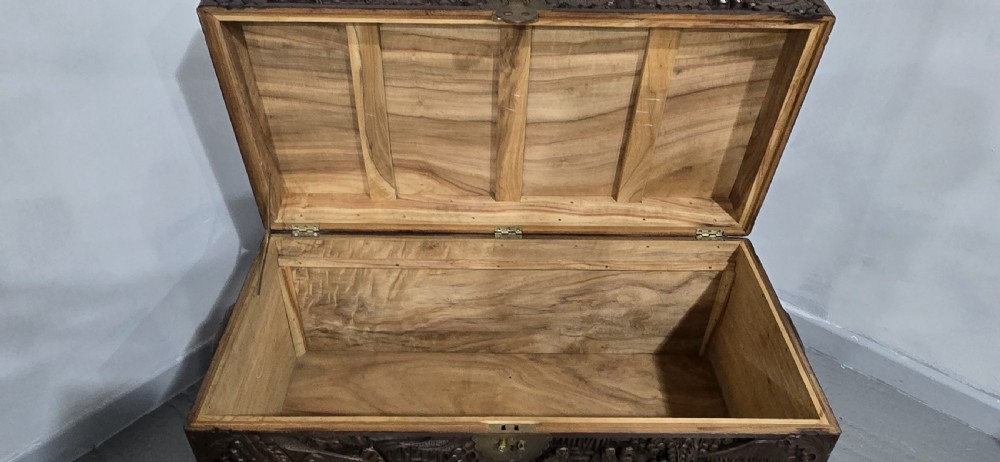 A rectangular dark timber storage chest featuring detailed relief carvings of figures and landscapes on all sides with a central brass clasp. View VIII