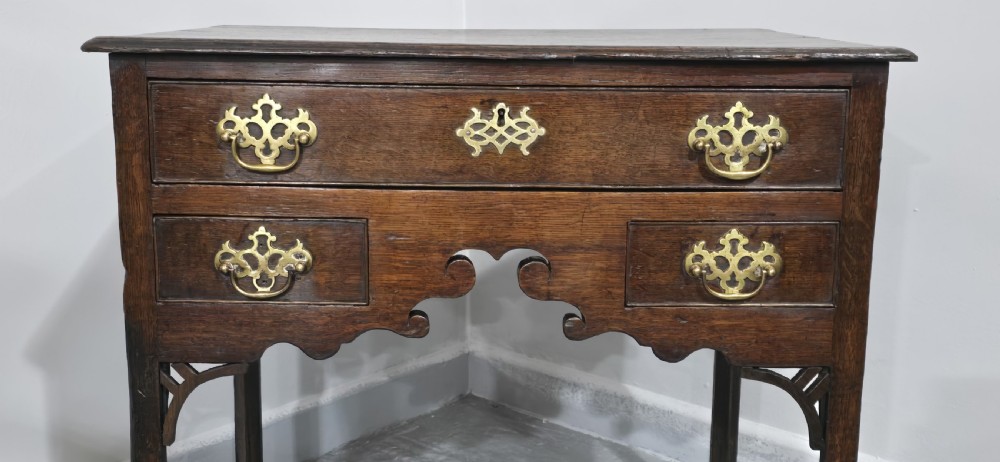 A dark oak 17th-century lowboy featuring three drawers with brass drop handles and a shaped kneehole apron set on straight square legs. View IV