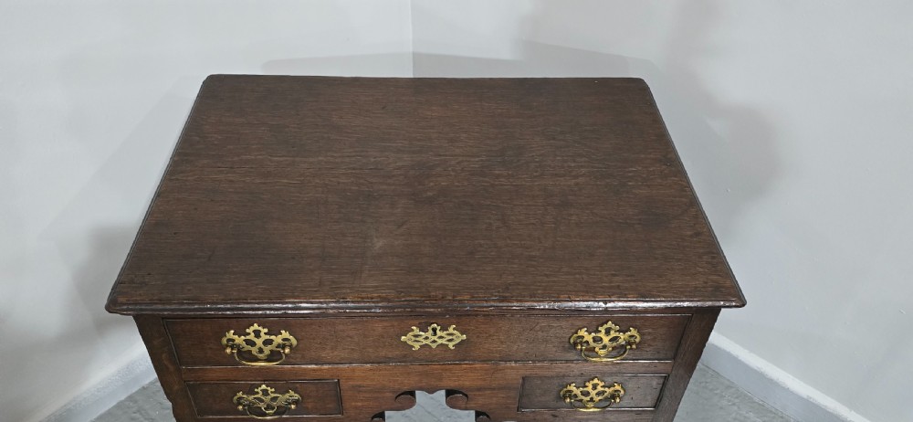 A dark oak 17th-century lowboy featuring three drawers with brass drop handles and a shaped kneehole apron set on straight square legs. View V