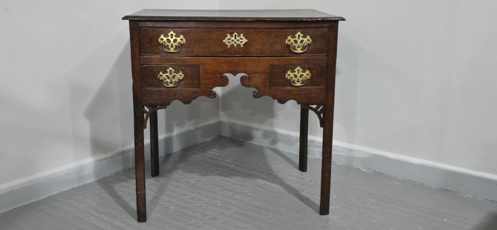 A dark oak 17th-century lowboy featuring three drawers with brass drop handles and a shaped kneehole apron set on straight square legs. View VI