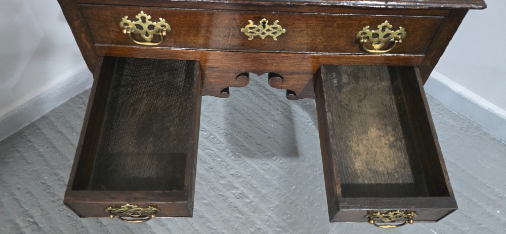 A dark oak 17th-century lowboy featuring three drawers with brass drop handles and a shaped kneehole apron set on straight square legs. View IX