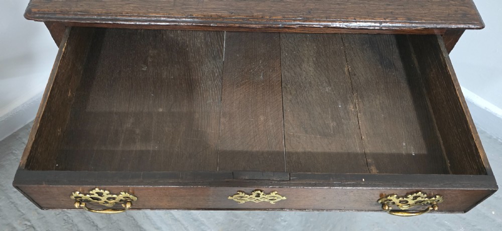 A dark oak 17th-century lowboy featuring three drawers with brass drop handles and a shaped kneehole apron set on straight square legs. View X