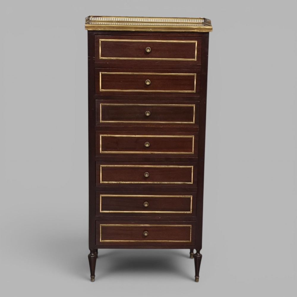 A tall, narrow dark mahogany chest of seven drawers with brass inlay banding, single knob handles, a brass gallery top, and turned tapered legs.