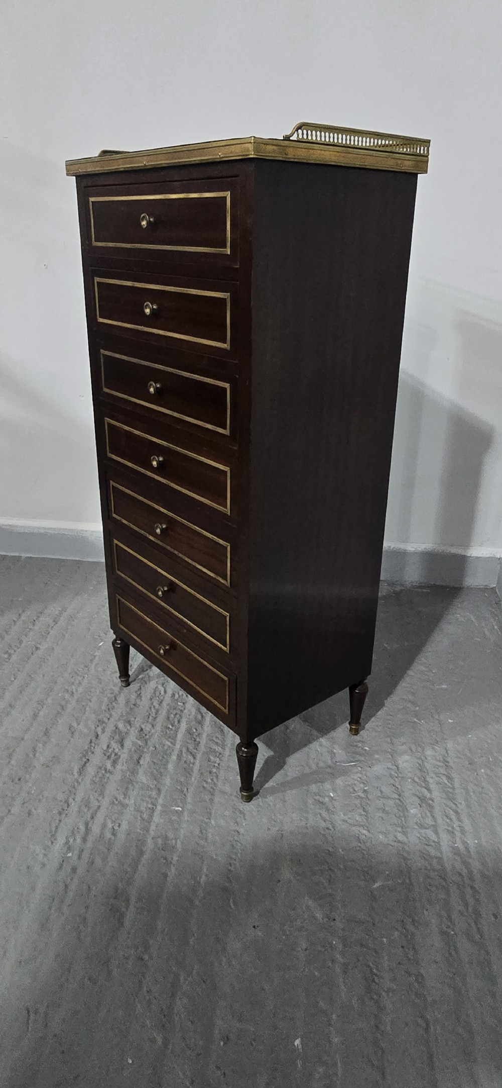 A tall, narrow dark mahogany chest of seven drawers with brass inlay banding, single knob handles, a brass gallery top, and turned tapered legs. View VI