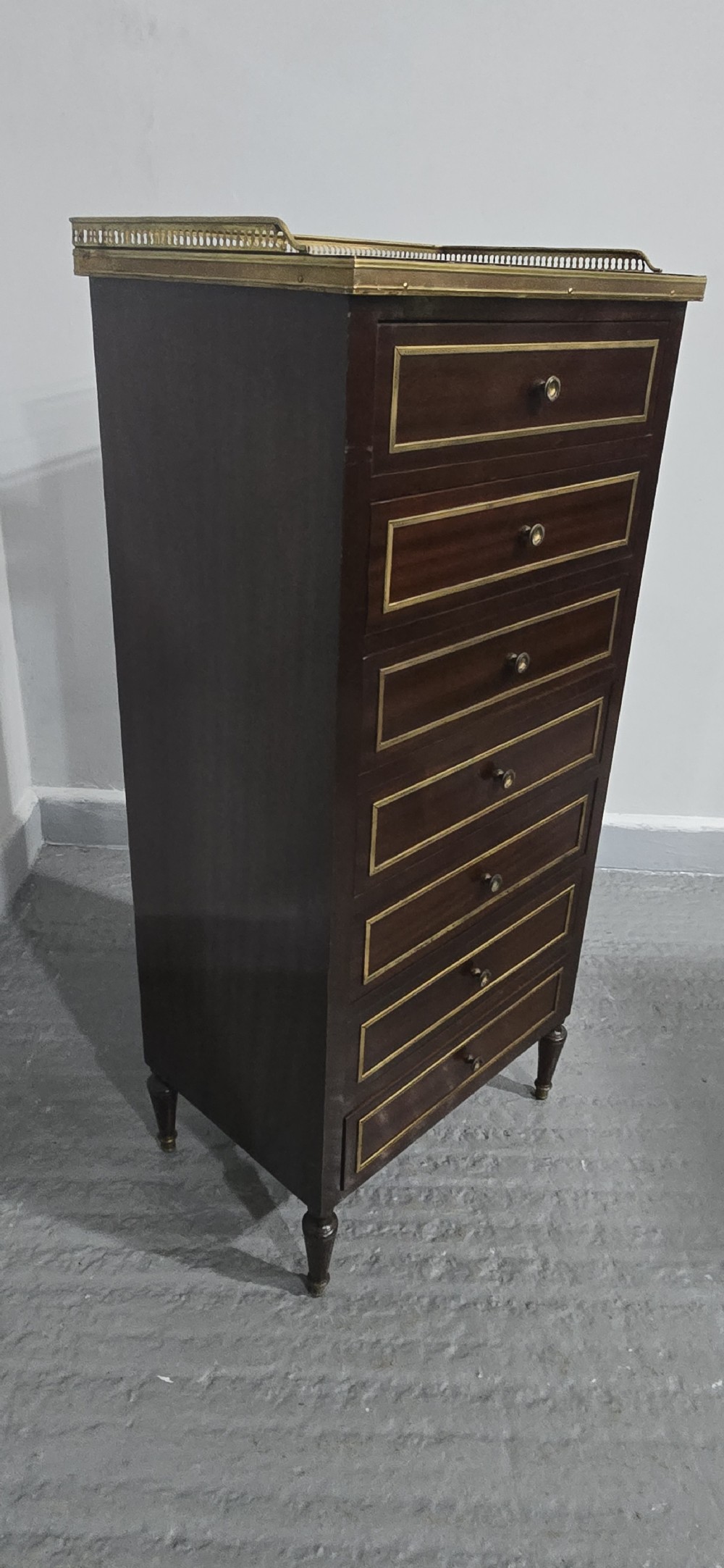 A tall, narrow dark mahogany chest of seven drawers with brass inlay banding, single knob handles, a brass gallery top, and turned tapered legs. View VII