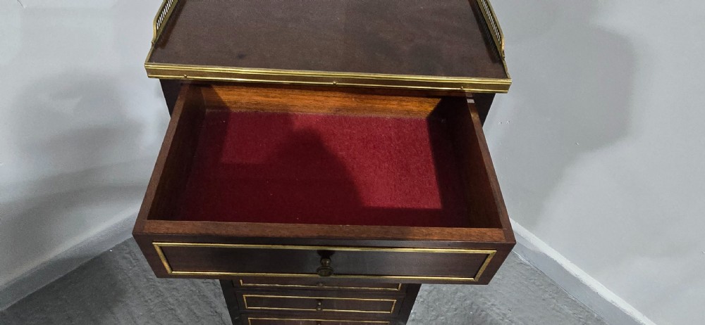 A tall, narrow dark mahogany chest of seven drawers with brass inlay banding, single knob handles, a brass gallery top, and turned tapered legs. View VIII