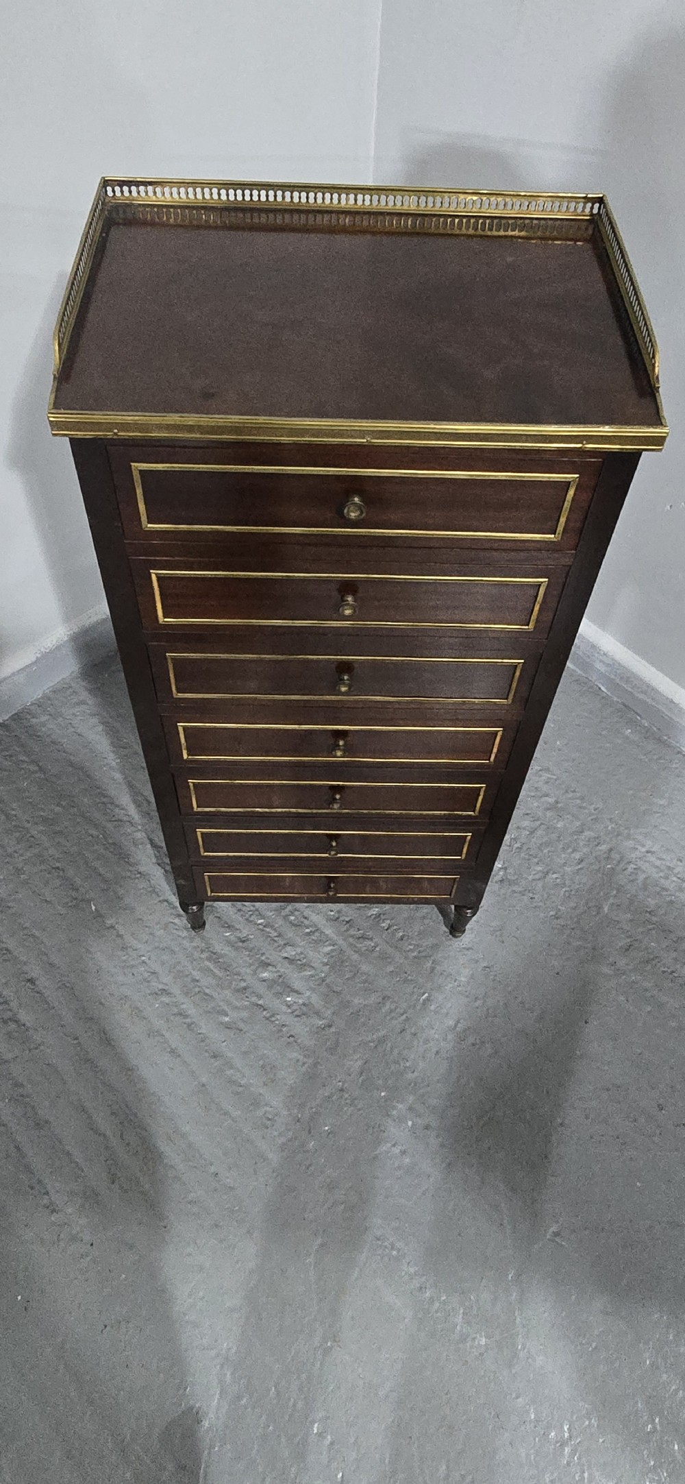 A tall, narrow dark mahogany chest of seven drawers with brass inlay banding, single knob handles, a brass gallery top, and turned tapered legs. View IX