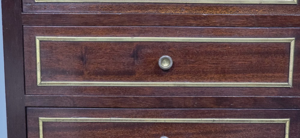 A tall, narrow dark mahogany chest of seven drawers with brass inlay banding, single knob handles, a brass gallery top, and turned tapered legs. View IV