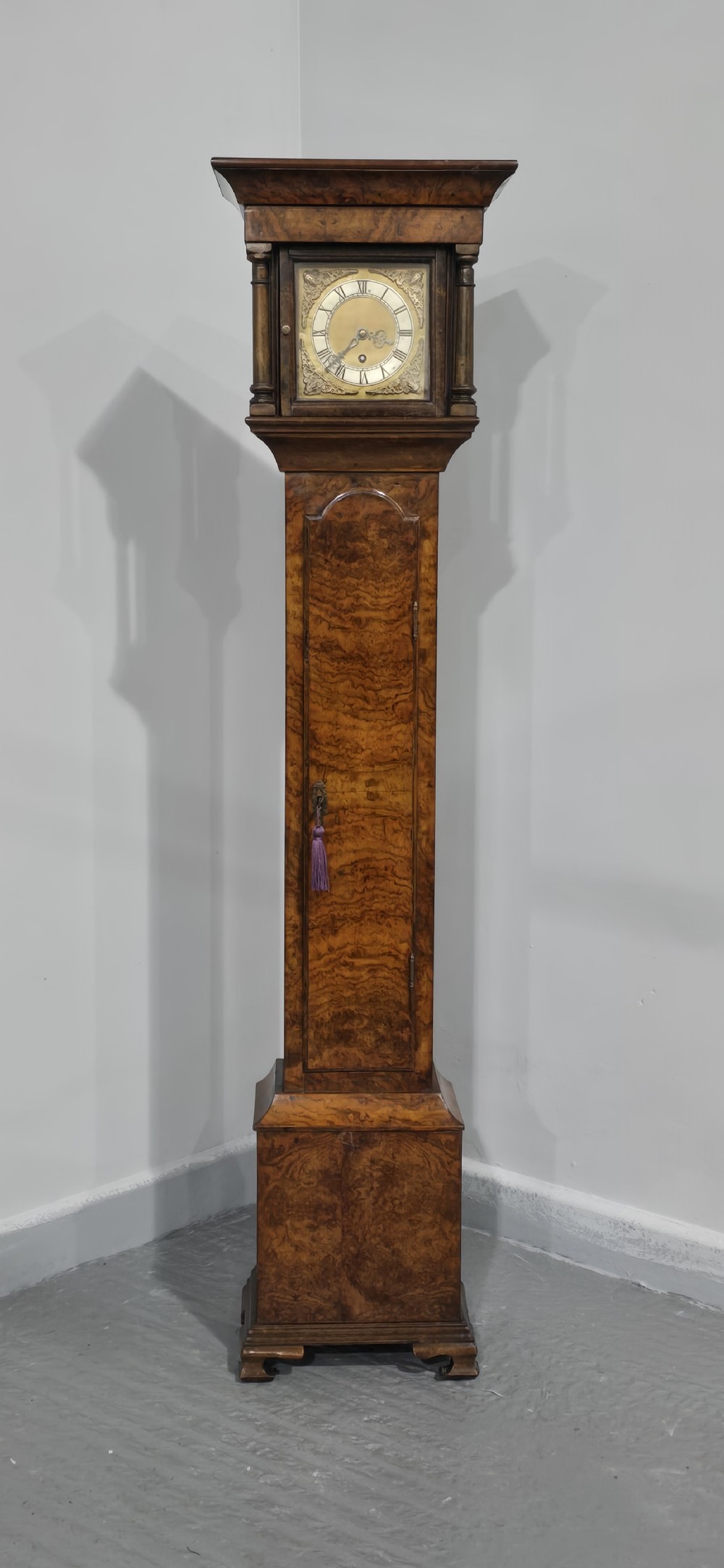 A slim floor-standing longcase clock crafted from figured burr walnut with a square brass dial and bracket feet. View III