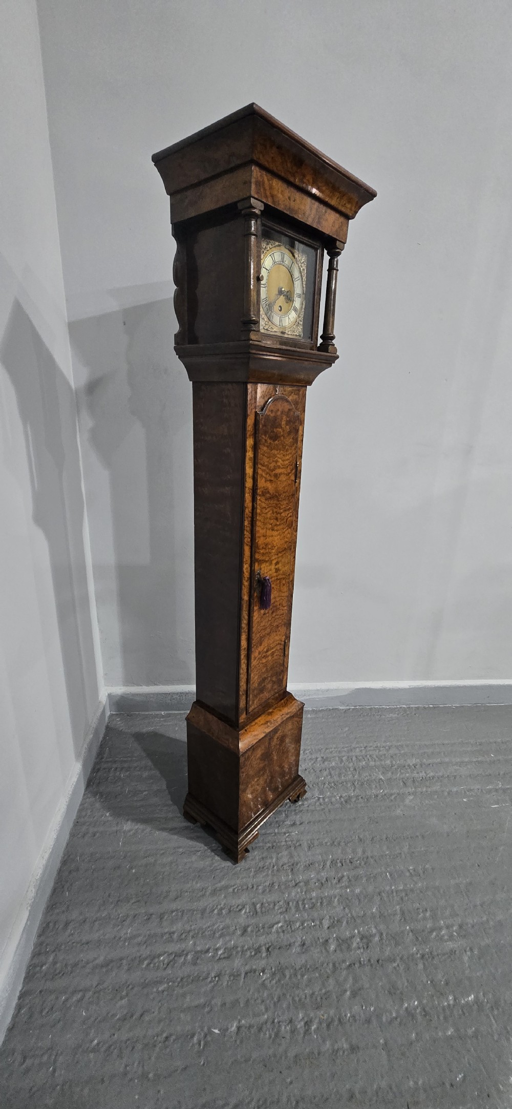 A slim floor-standing longcase clock crafted from figured burr walnut with a square brass dial and bracket feet. View XIV