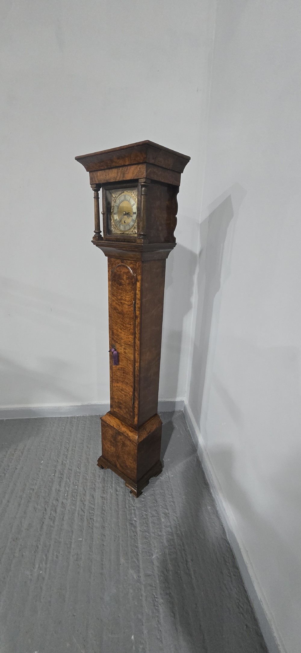 A slim floor-standing longcase clock crafted from figured burr walnut with a square brass dial and bracket feet. View XV