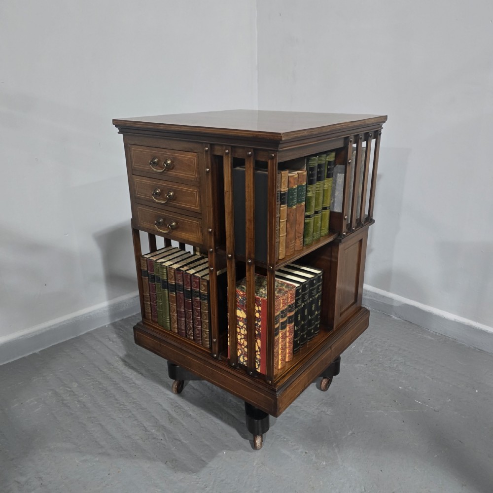 An Edwardian inlaid mahogany revolving bookcase featuring three small side drawers with brass drop handles, vertical timber slats, and metal castors on the base. View III