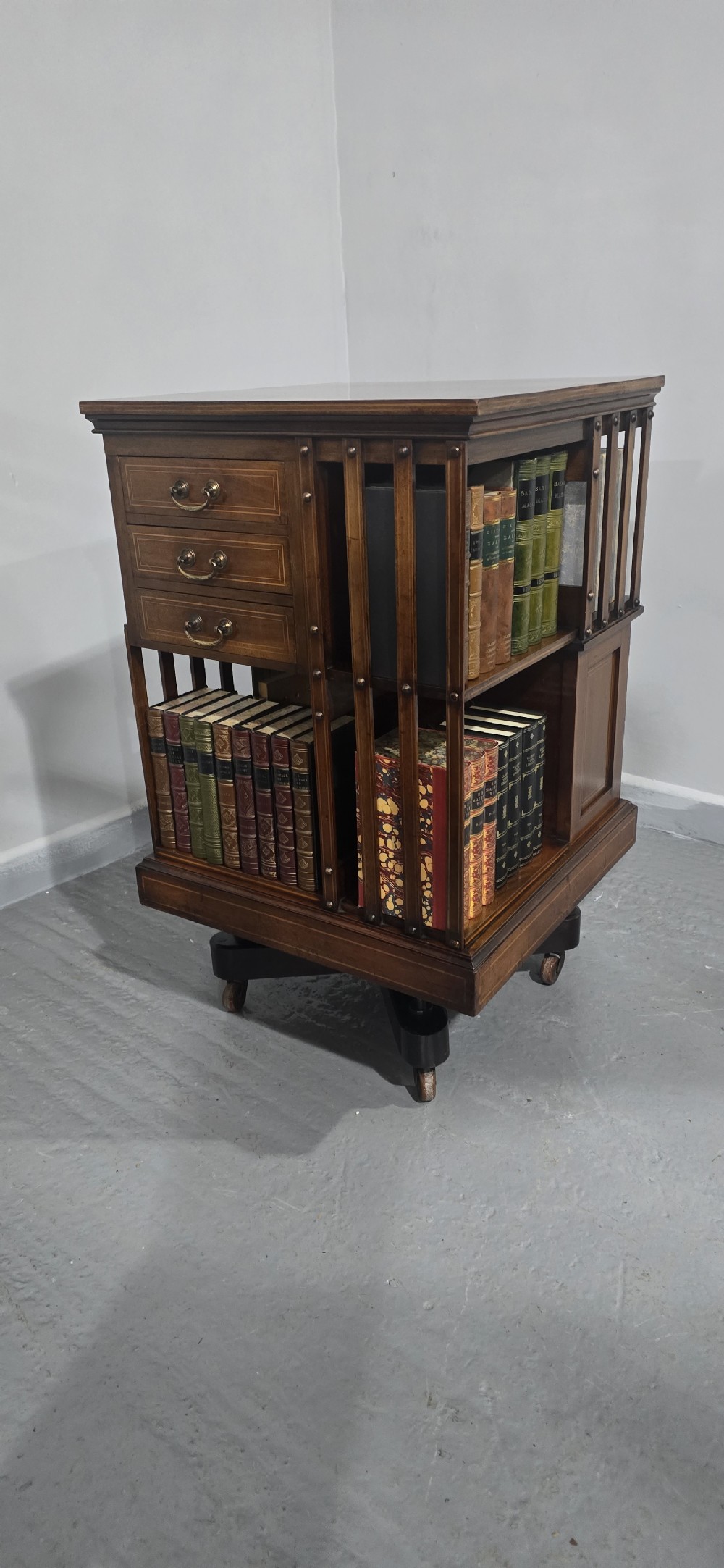 An Edwardian inlaid mahogany revolving bookcase featuring three small side drawers with brass drop handles, vertical timber slats, and metal castors on the base. View V