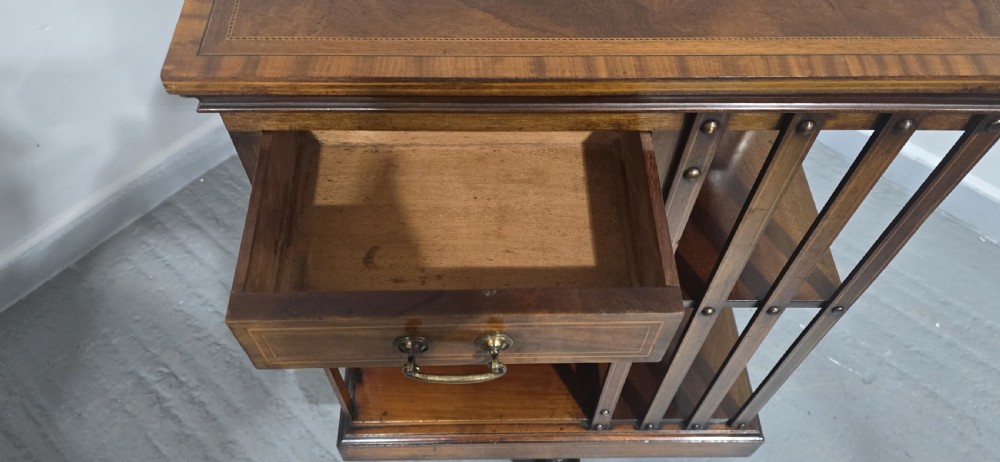 An Edwardian inlaid mahogany revolving bookcase featuring three small side drawers with brass drop handles, vertical timber slats, and metal castors on the base. View X