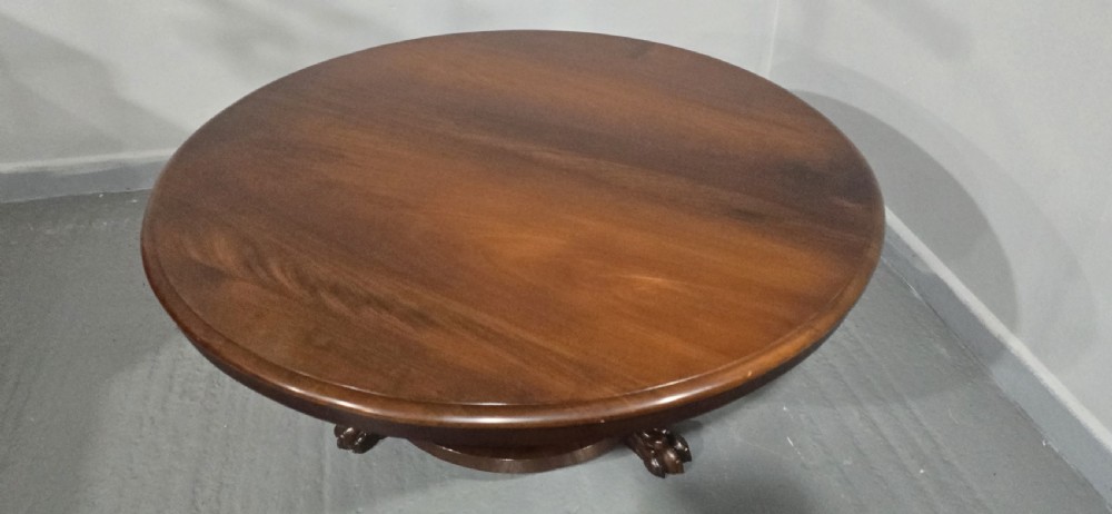 A low, circular mahogany coffee table featuring a smooth polished top supported by a central turned pedestal with carved claw feet. View VII