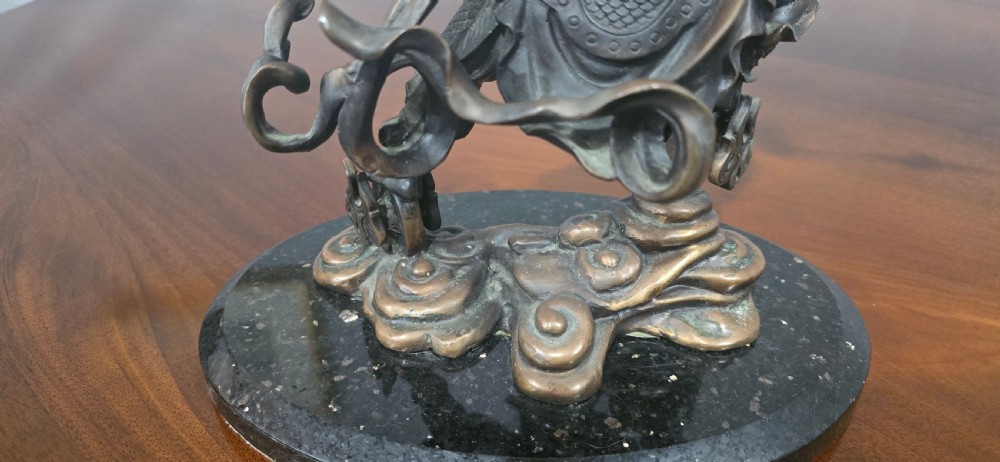 A detailed bronze sculpture depicting the Chinese mythological figure Ne Zha holding a spear, mounted on a circular black marble base. View X