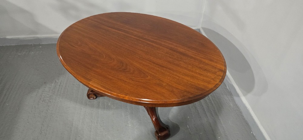 An oval mahogany coffee table featuring a polished surface, a central turned pedestal, and four carved cabriole legs terminating in scroll feet. View VII