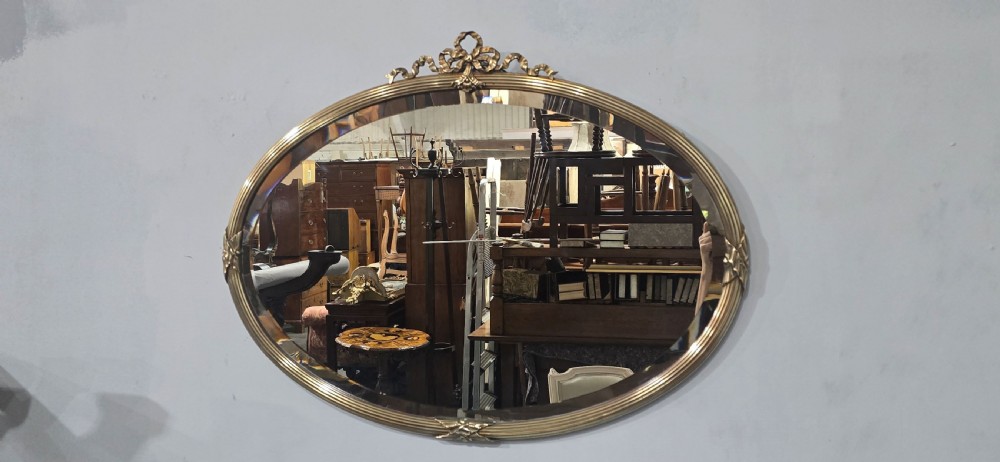 A horizontal oval Victorian brass wall mirror featuring a decorative ribbon-and-bow crest and bevelled mirror plate. View II