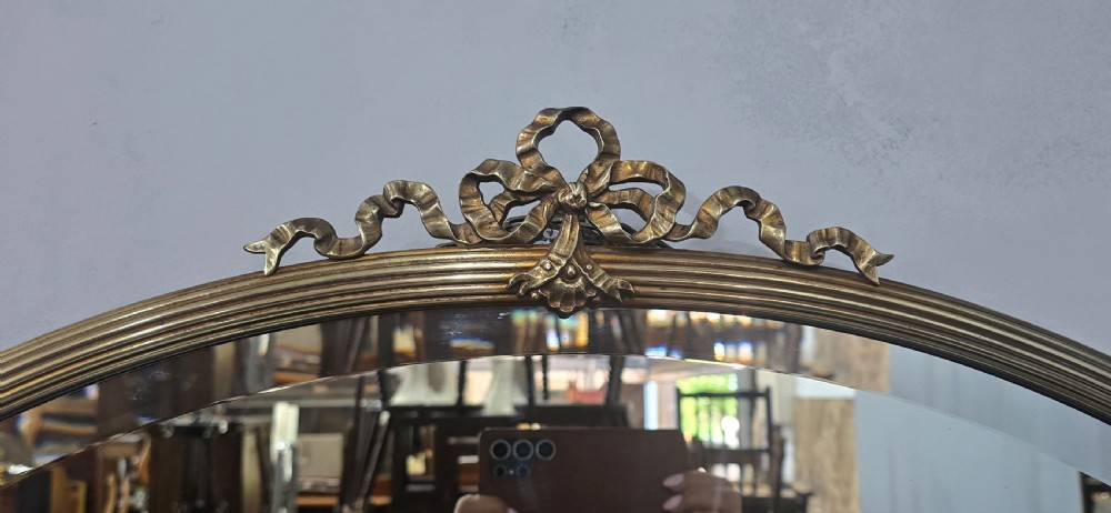 A horizontal oval Victorian brass wall mirror featuring a decorative ribbon-and-bow crest and bevelled mirror plate. View IV