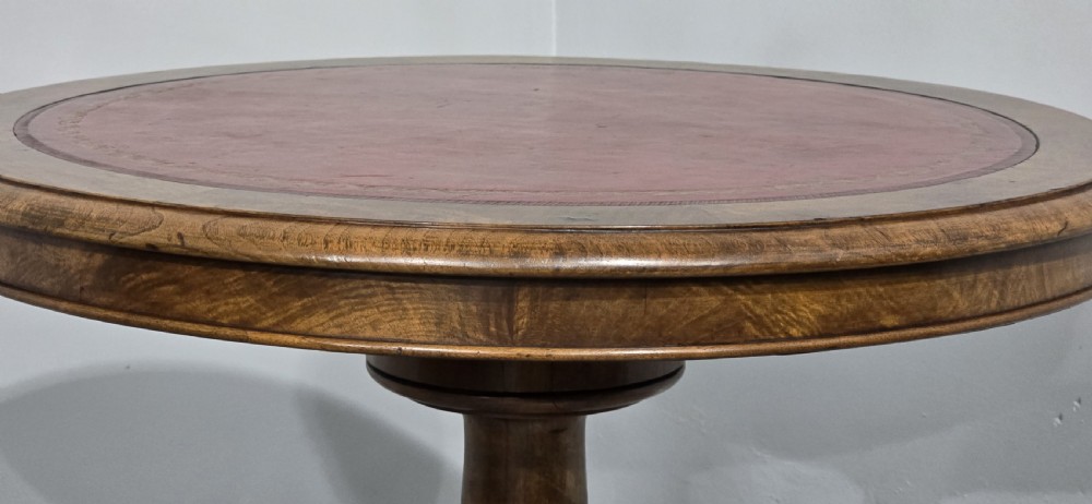 A circular Victorian walnut library table featuring a reddish-brown leather inset top, turned central pedestal stem, and carved tripod legs with scroll feet. View VII