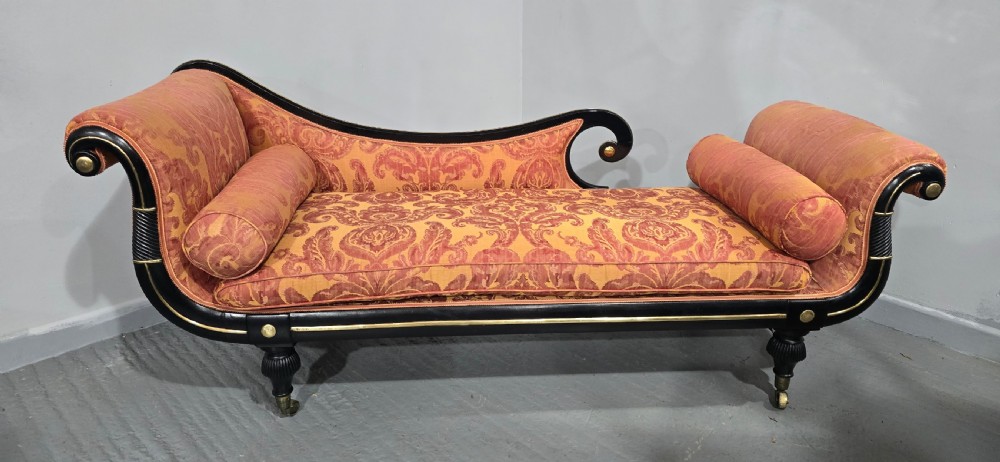 An ebonised Regency chaise longue sofa with gilt details, scrolled ends, turned legs on brass castors, and patterned terracotta damask upholstery with matching bolster cushions. View IV