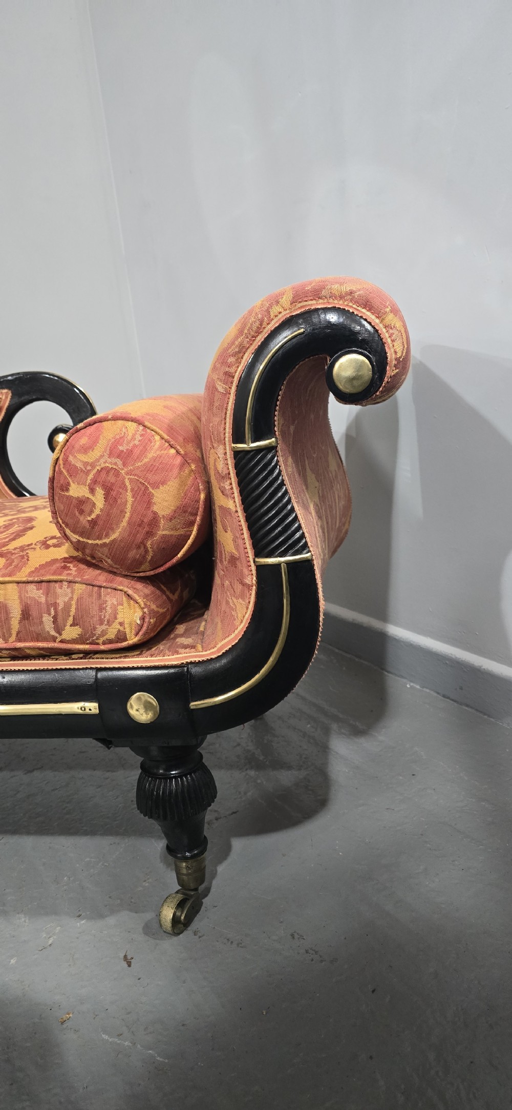 An ebonised Regency chaise longue sofa with gilt details, scrolled ends, turned legs on brass castors, and patterned terracotta damask upholstery with matching bolster cushions. View V