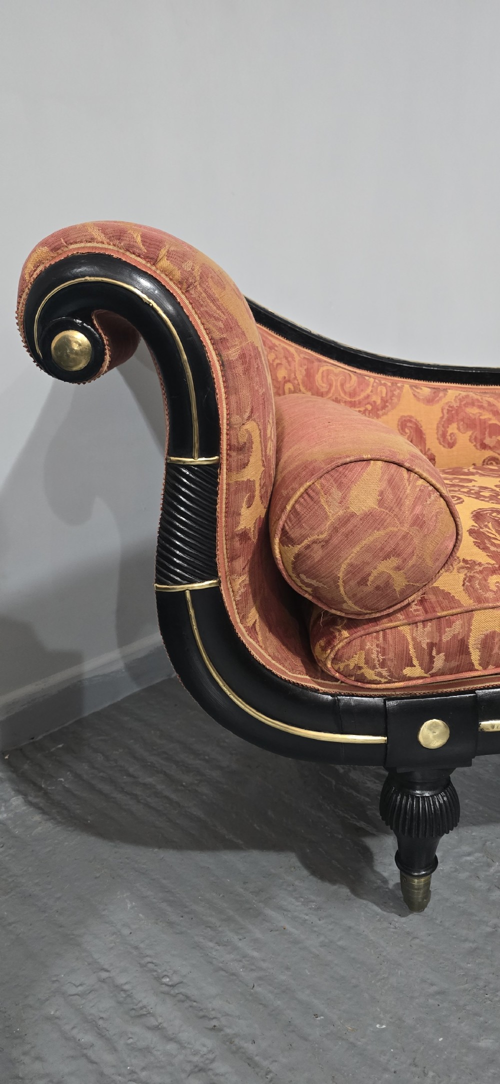 An ebonised Regency chaise longue sofa with gilt details, scrolled ends, turned legs on brass castors, and patterned terracotta damask upholstery with matching bolster cushions. View VI