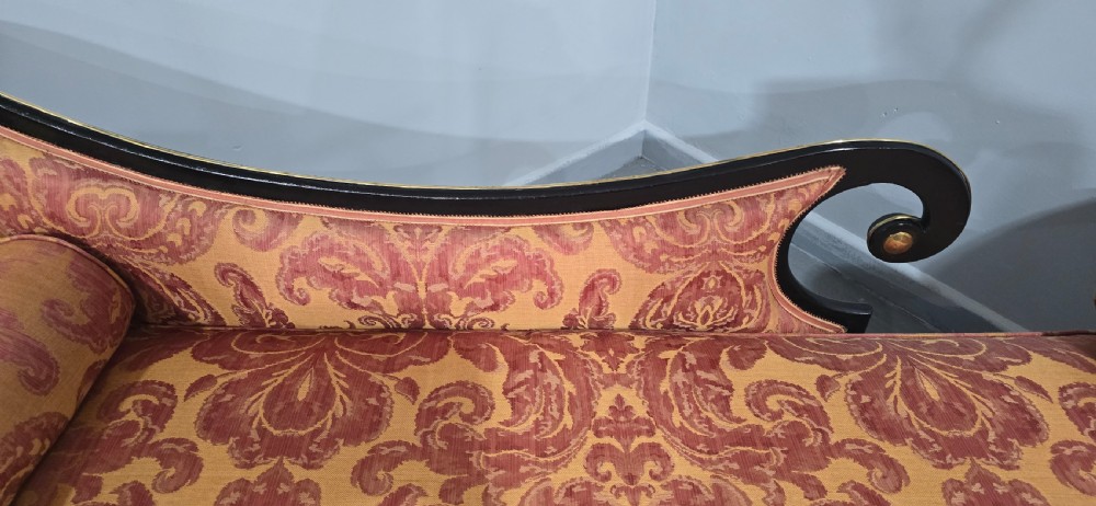 An ebonised Regency chaise longue sofa with gilt details, scrolled ends, turned legs on brass castors, and patterned terracotta damask upholstery with matching bolster cushions. View VII