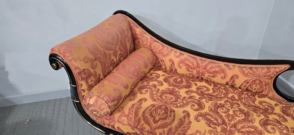 An ebonised Regency chaise longue sofa with gilt details, scrolled ends, turned legs on brass castors, and patterned terracotta damask upholstery with matching bolster cushions. View IX