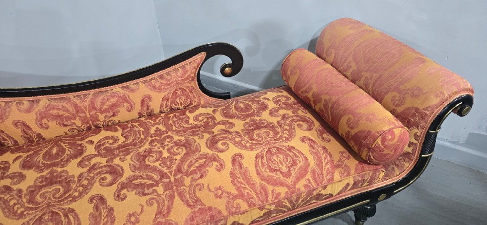 An ebonised Regency chaise longue sofa with gilt details, scrolled ends, turned legs on brass castors, and patterned terracotta damask upholstery with matching bolster cushions. View X