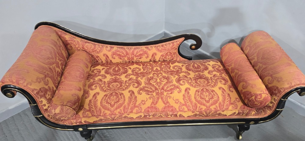 An ebonised Regency chaise longue sofa with gilt details, scrolled ends, turned legs on brass castors, and patterned terracotta damask upholstery with matching bolster cushions. View XI