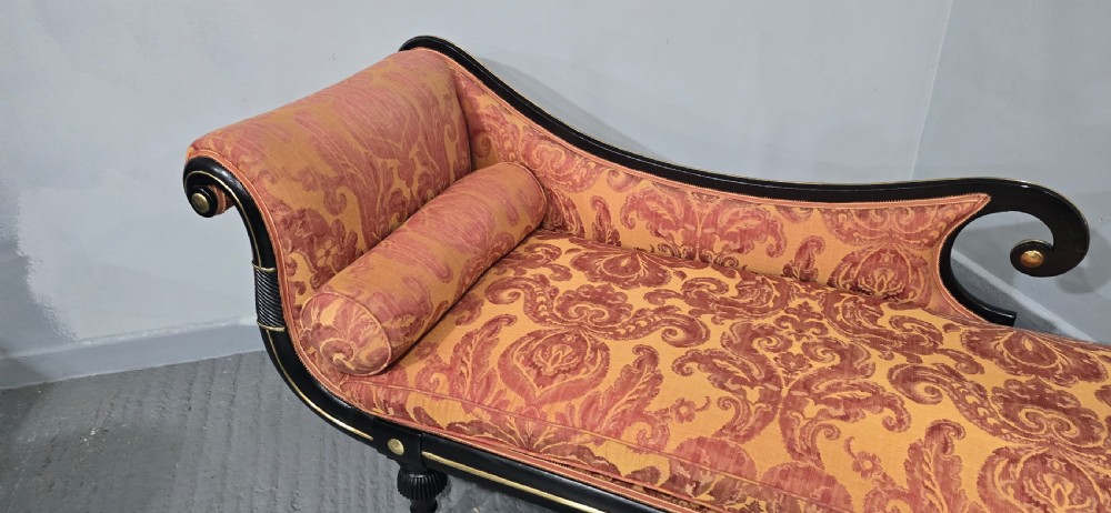 An ebonised Regency chaise longue sofa with gilt details, scrolled ends, turned legs on brass castors, and patterned terracotta damask upholstery with matching bolster cushions. View XII