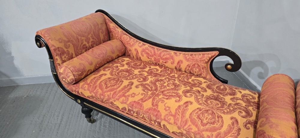 An ebonised Regency chaise longue sofa with gilt details, scrolled ends, turned legs on brass castors, and patterned terracotta damask upholstery with matching bolster cushions. View XIII