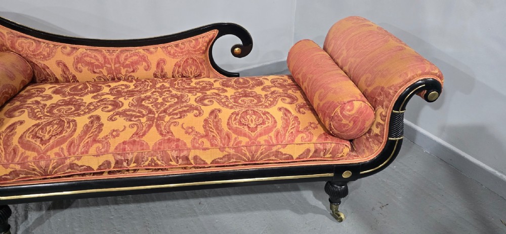 An ebonised Regency chaise longue sofa with gilt details, scrolled ends, turned legs on brass castors, and patterned terracotta damask upholstery with matching bolster cushions. View XIV