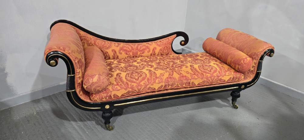 An ebonised Regency chaise longue sofa with gilt details, scrolled ends, turned legs on brass castors, and patterned terracotta damask upholstery with matching bolster cushions. View XV