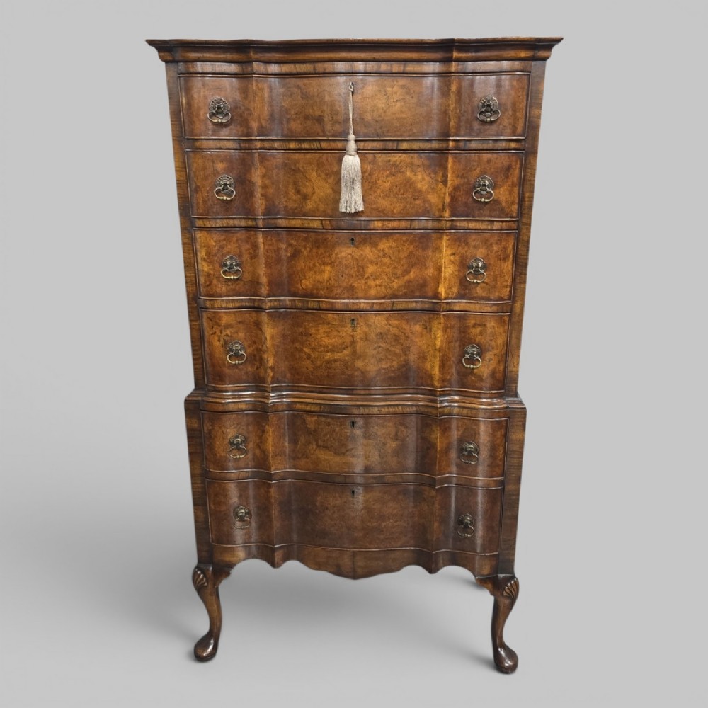 A tall Queen Anne burr walnut chest on stand featuring a serpentine contoured front with six drawers, brass drop handles, and carved cabriole legs.