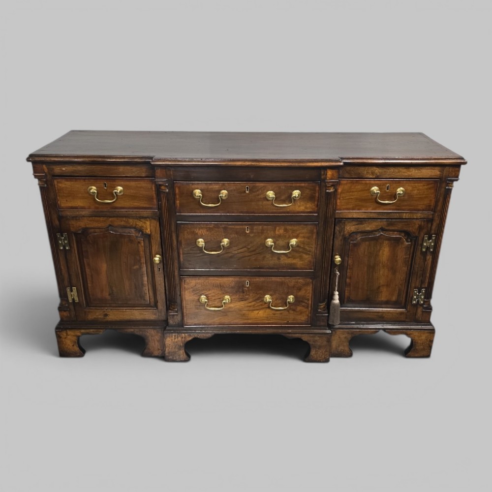 A dark oak Georgian breakfront dresser base featuring brass swan-neck handles, three central drawers, and two outer cupboard doors set on bracket feet.