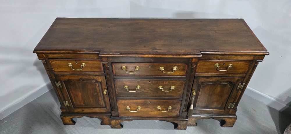 A dark oak Georgian breakfront dresser base featuring brass swan-neck handles, three central drawers, and two outer cupboard doors set on bracket feet. View IV
