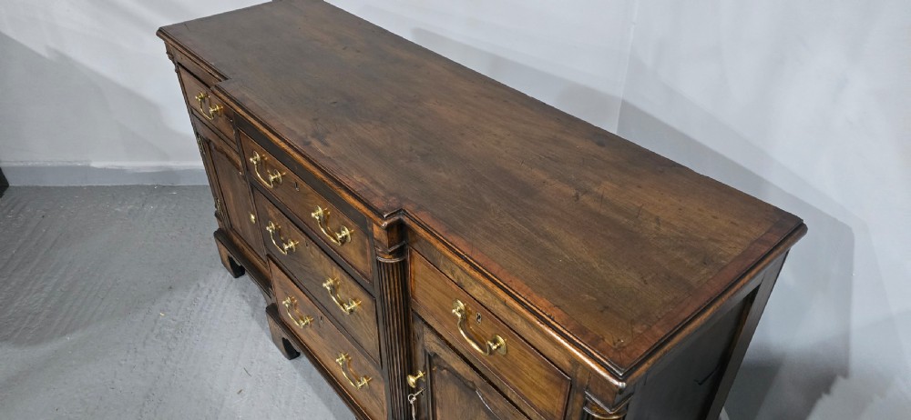 A dark oak Georgian breakfront dresser base featuring brass swan-neck handles, three central drawers, and two outer cupboard doors set on bracket feet. View V